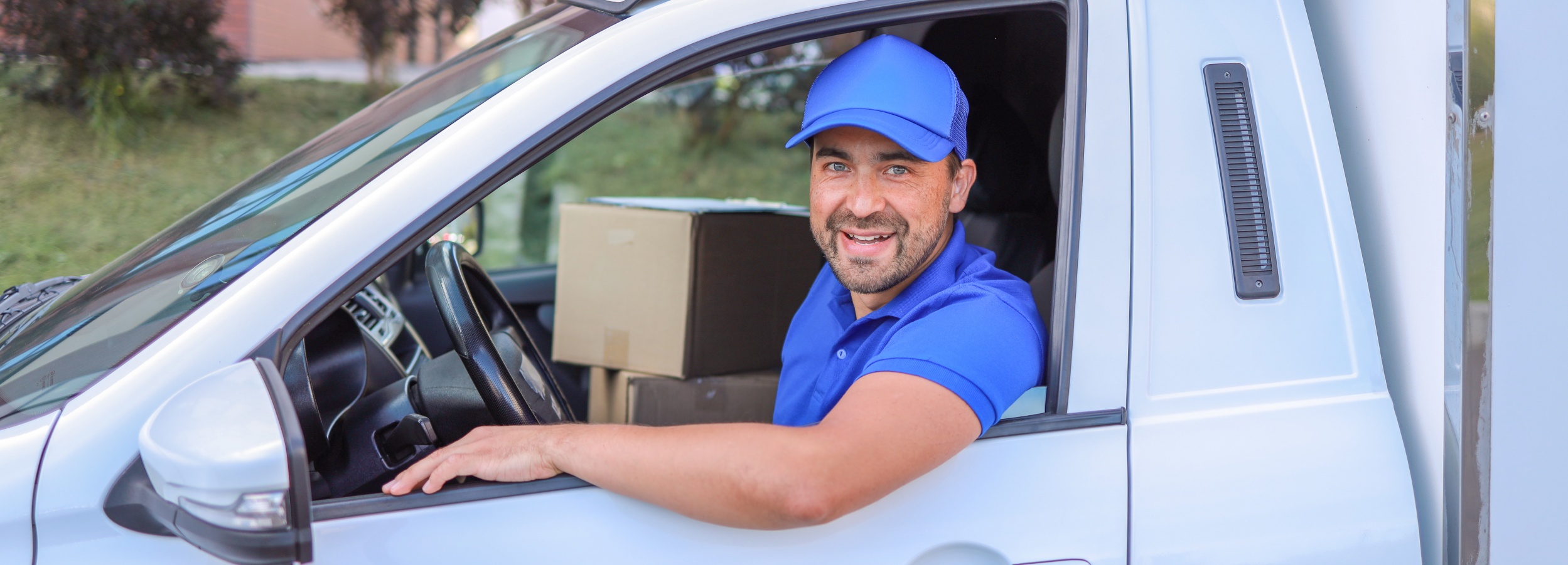 Ein freundlicher Lieferfahrer in blauer Uniform sitzt lächelnd in einem weißen Lieferwagen, dessen Innenraum mit Kartons gefüllt ist.

Das Bild zeigt einen Lieferfahrer, der in einem weißen Lieferwagen sitzt. Der Mann trägt eine blaue Uniform und eine blaue Kappe. Er lächelt in die Kamera. Im Hintergrund des Vans befinden sich mehrere Kartons, die ordentlich gestapelt sind. Der Fahrer hat eine Hand am Lenkrad und schaut freundlich aus dem Fenster. Die Atmosphäre wirkt positiv und einladend. Der Hintergrund ist unscharf, wodurch der Fokus auf dem Fahrer und dem Lieferwagen liegt. Das Bild vermittelt einen Eindruck von Zuverlässigkeit und Kundenservice.