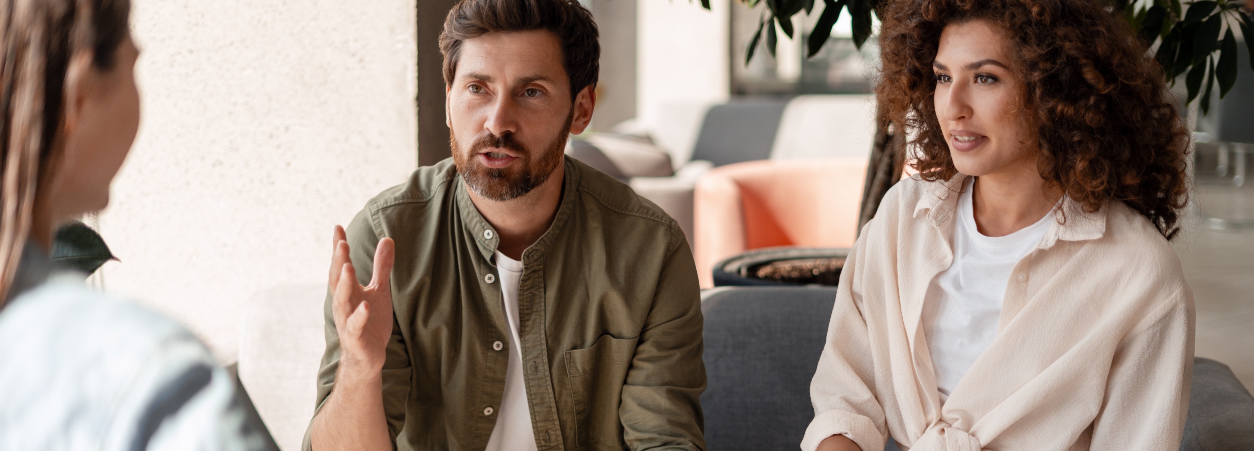 Das Bild zeigt ein lebhaftes Gespräch zwischen drei Personen in einem modernen, hellen Raum.

Das Bild zeigt drei Personen, möglicherweise in einem Büro oder einem informellen Besprechungsraum. Ein Mann mit Bart und dunklem Haar trägt ein olivgrünes Hemd und gestikuliert mit seiner Hand, während er spricht. Auf der rechten Seite sitzt eine Frau mit lockigem Haar, die ein helles Hemd über einem weißen T-Shirt trägt und aufmerksam zuhört. Die dritte Person ist nur teilweise im Bild zu sehen. Im Hintergrund sind moderne Möbel, darunter graue und pfirsichfarbene Sitzelemente, sowie Pflanzen zu erkennen, die für eine angenehme Atmosphäre sorgen. Das Licht ist hell und natürlich, was die Stimmung des Bildes freundlich und einladend erscheinen lässt. Die Szene wirkt wie ein offenes, kollaboratives Gespräch.
