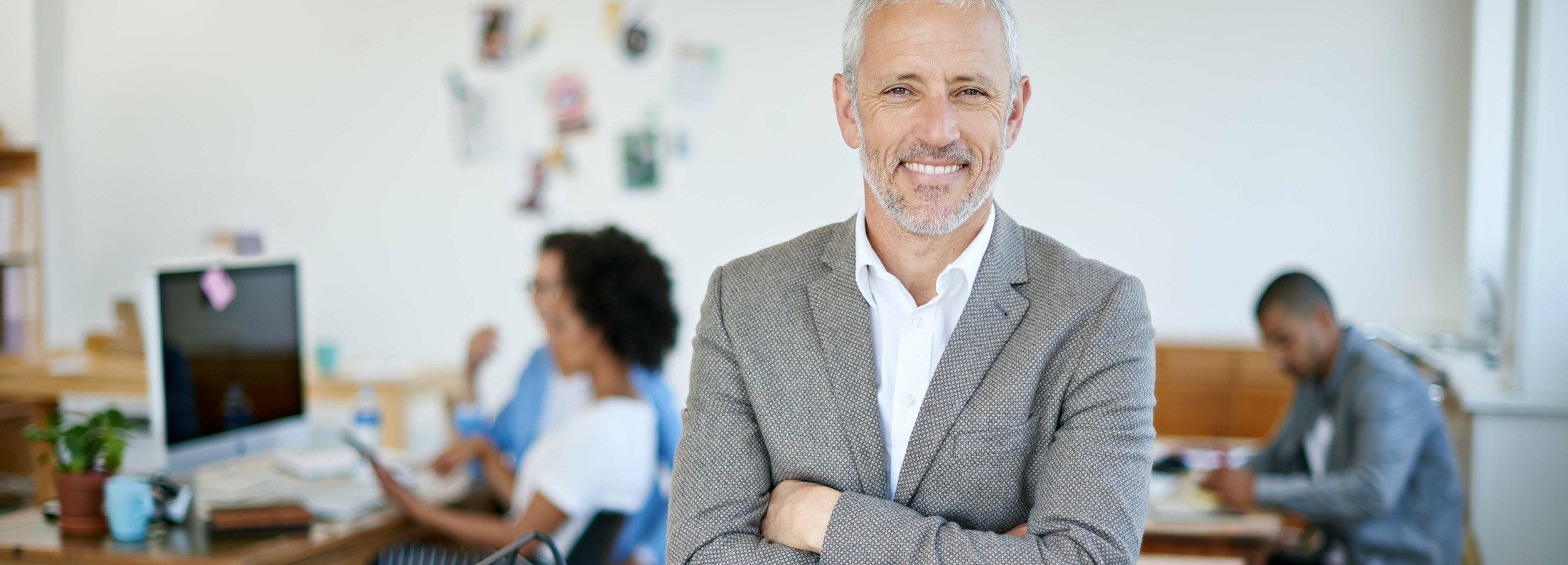 Ein gutaussehender, selbstbewusster Geschäftsmann steht mit verschränkten Armen im Vordergrund, während im Hintergrund ein belebtes Büro mit arbeitenden Kollegen andeutet.

Das Bild zeigt den Fokus auf einen Mann mittleren Alters mit grauen Haaren und Bart, der in die Kamera lächelt. Er trägt ein helles Hemd und darüber ein anthrazitfarbenes Sakko, seine Arme sind vor der Brust verschränkt, was ihm eine selbstsichere Haltung verleiht. Im Hintergrund sind zwei weitere Personen an Schreibtischen zu sehen, die beschäftigt mit Computern und Tablets arbeiten. Das Büro scheint hell und modern zu sein, mit weißen Wänden, an denen einige Dekorationen angebracht sind, und hölzernen Schreibtischen. Die allgemeine Atmosphäre vermittelt Professionalität und eine positive Arbeitsumgebung.