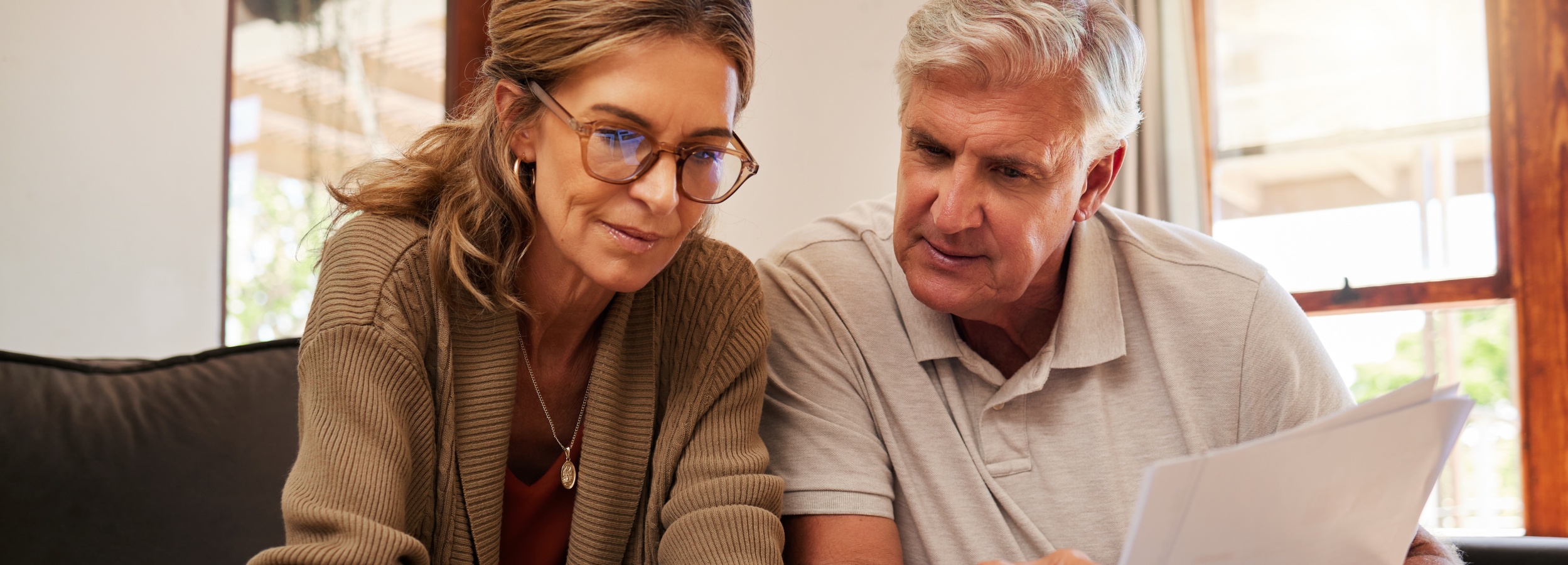 Ein älteres Paar sitzt nah beieinander und bespricht konzentriert einige Dokumente, was auf eine gemeinsame finanziellen oder rechtlichen Angelegenheit hindeutet.

Auf dem Bild sind ein älterer Mann und eine ältere Frau zu sehen, die nebeneinander auf einem Sofa oder einer Couch sitzen. Sie sind beide im Profil oder halb von der Seite zu sehen und blicken auf Papiere, die vermutlich in ihren Händen gehalten werden, obwohl diese nicht eindeutig erkennbar sind. Der Mann hat graue Haare, trägt ein hellgraues Poloshirt und wirkt aufmerksam und nachdenklich. Die Frau hat hellbraune, schulterlange Haare, die locker fallen, und trägt eine braune Strickjacke über einem orangen Oberteil. Sie trägt eine auffällige Brille mit braunem Rahmen und eine Halskette mit einem ovalen Anhänger. Ihr Blick ist ebenfalls auf die Dokumente gerichtet, und sie scheint ihrerseits konzentriert zu sein. Der Hintergrund ist unscharf und deutet auf ein Wohnzimmer hin, mit einem Fenster, durch das Tageslicht fällt und das eine leicht unscharfe, grüne Vegetation im Außenbereich erahnen lässt. Die Atmosphäre des Bildes ist ruhig, konzentriert und intim. Die Stimmung wirkt seriös, aber nicht bedrückend, eher wie ein gemeinsames Bewältigen einer wichtigen Aufgabe. Es strahlt Vertrauen und Partnerschaft aus, da beide sich der gleichen Sache widmen.