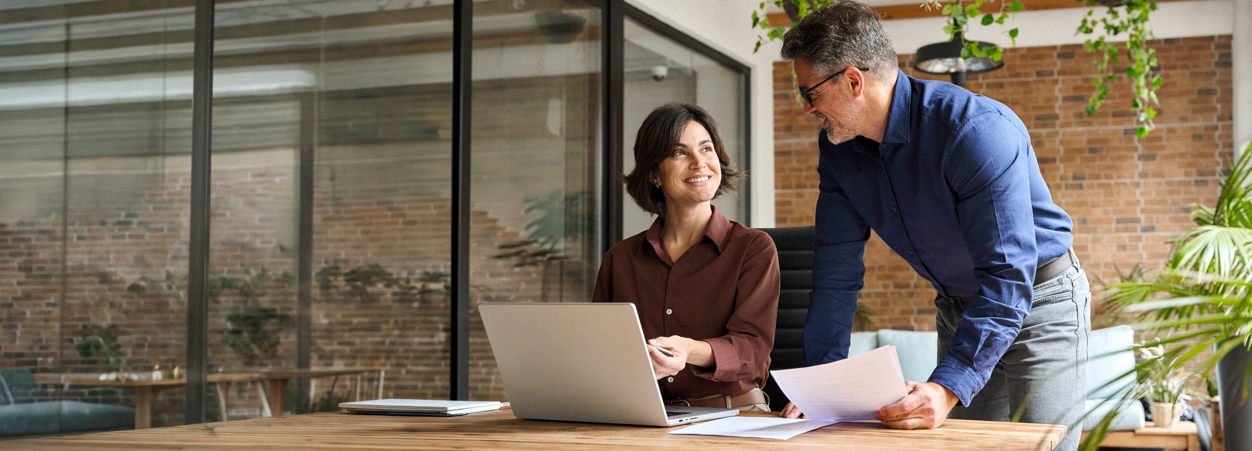 Zwei Kollegen arbeiten im Büro zusammen und besprechen eine Angelegenheit.

Im Bild sind zwei Personen zu sehen, die in einem modernen Büro arbeiten. Im Vordergrund sitzt eine Frau an einem Holztisch vor einem geöffneten Laptop und lächelt, während sie mit einem Stift auf den Bildschirm zeigt. Sie hat dunkle, schulterlange Haare und trägt eine braune Bluse. Ihr gegenüber steht ein Mann, der sich leicht über den Tisch beugt und ihr ein Dokument in die Hand drückt, während er sie anlächelt. Er hat kurze graue Haare und trägt eine blaue Hemdbluse und Jeans. Der Hintergrund ist durch eine Glaswand mit Blick auf eine Ziegelwand und Pflanzen getrennt. Die Beleuchtung ist weich und warm, was eine angenehme und produktive Atmosphäre schafft. Die Stimmung ist positiv und kollaborativ, was darauf hindeutet, dass die beiden eine gute Arbeitsbeziehung haben und an einer gemeinsamen Aufgabe arbeiten.