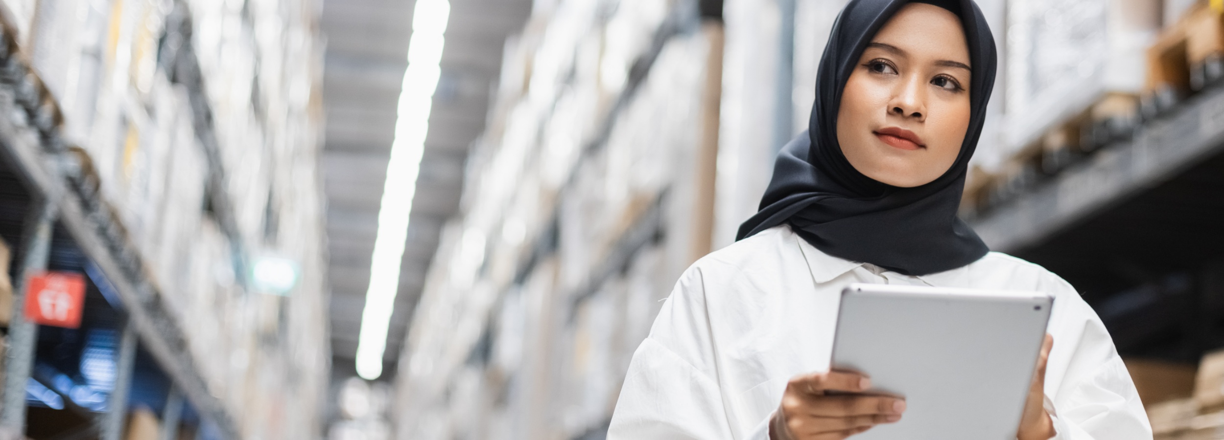 Das Bild zeigt eine junge Frau in einem Lager, die ein Tablet in der Hand hält.

Das Bild ist ein Panoramabild, das eine Nahaufnahme einer jungen Frau zeigt, die eine schwarze Hijab und ein weißes Hemd trägt. Sie blickt nach rechts und hält ein silbernes Tablet in ihren Händen. Hinter ihr erstreckt sich ein breiter Gang eines Lagers, flankiert von hohen Regalen, die mit zahlreichen Kartons gefüllt sind. Die Regale sind in einer symmetrischen Anordnung angeordnet und erzeugen eine tiefe Perspektive. Über den Regalen sind helle Leuchtstoffröhren angebracht, die den Raum gleichmäßig ausleuchten und für eine klare, fast klinische Atmosphäre sorgen. Die Beleuchtung ist hell und diffus, was auf eine professionelle und organisierte Umgebung hindeutet. Die Stimmung des Bildes ist ruhig und konzentriert. Die Frau scheint in ihre Aufgabe vertieft zu sein, was sie zu einer aktiven und kompetenten Person in dieser Arbeitsumgebung macht. Die weiche Schärfe im Hintergrund lenkt den Fokus auf die Frau und ihre Tätigkeit, während die gestaffelten Regale eine klare räumliche Tiefe vermitteln.